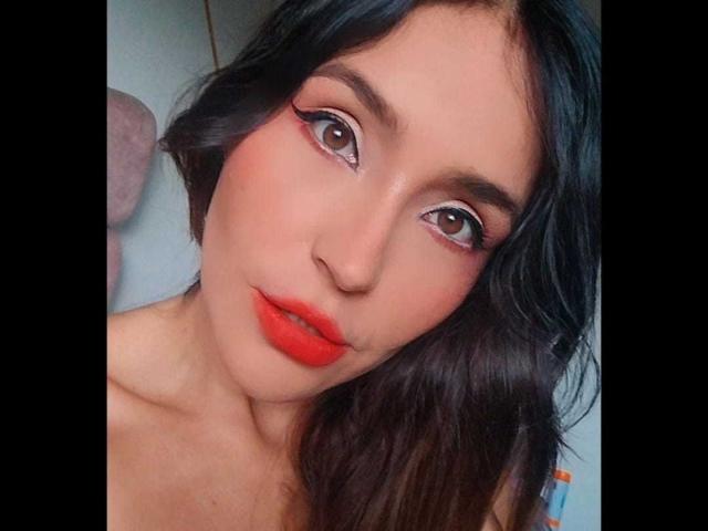 Il viso di MindyLove, con rossetto rosso, capelli neri ondulati e un trucco per gli occhi marcato, mostra i suoi occhi marroni e le labbra leggermente aperte in un selfie. Il suo seno non è visibile..