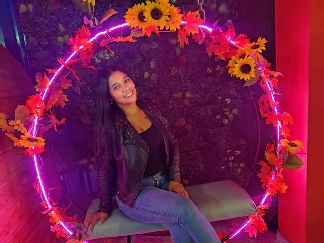 PauletteLoveX, com cabelos pretos longos, jaqueta de couro, blusa preta e jeans azuis, sentada em um balanço em forma de anel de cor roxa neon, com girassóis, sorrindo. Suas nádegas estão ligeiramente visíveis..