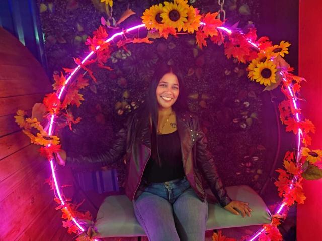 PauletteLoveX est assise sur une balançoire en forme de cercle rose fluo, décorée de tournesols, et porte un haut noir, un jean bleu et une veste en cuir. Elle sourit, avec ses fesses légèrement relevées..