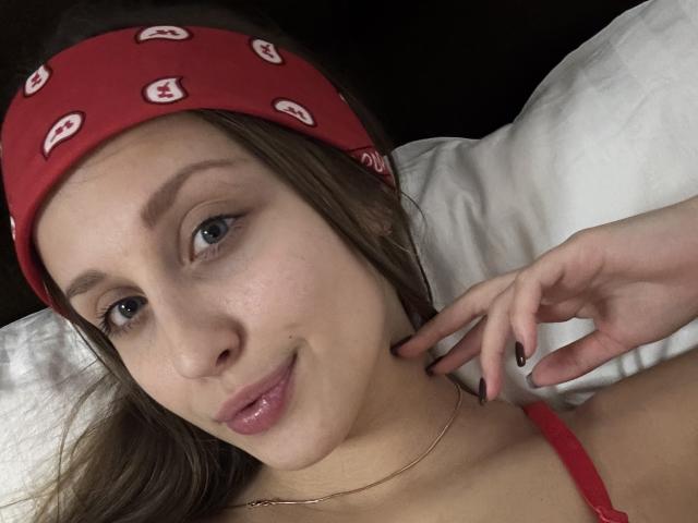 TrissyTerry, avec un bandana rouge, des cheveux bruns clairs, des yeux bleus et un léger sourire, portant une bretelle rouge, allongée sur des oreillers blancs, touchant son cou..