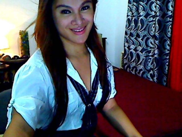 Une jeune femme philippine attrayante, avec de longs cheveux bruns, un chemisier blanc, une robe noire à lacets, souriante, montrant ses seins, assise sur un canapé rouge, avec des rideaux à motifs en arrière-plan..