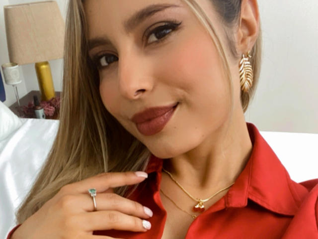Amara Ricci mit blonden Haaren, rotem Hemd, goldenen Ohrringen, einem Ring, einer Halskette und Lippenstift, lächelt in die Kamera, zeigt die Zunge und hält den Kragen des Hemdes. Brüste nicht sichtbar. Hintern nicht sichtbar. Vulva nicht sichtbar..