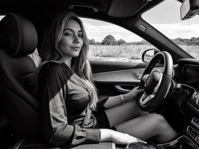 Sofie White met lang haar, zittend in een auto, een shirt aan, de stuurwiel vasthoudend, naar de camera kijkend, haar borsten onthullend, op een zwart-wit foto..