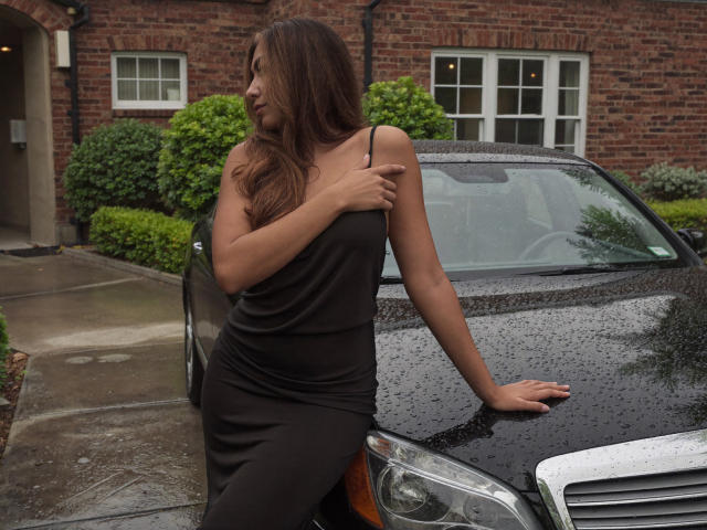SofieWhite with long brown hair, black dress, and small tits, leaning on a wet black car, right arm on roof, left hand on chest, looking to the side.