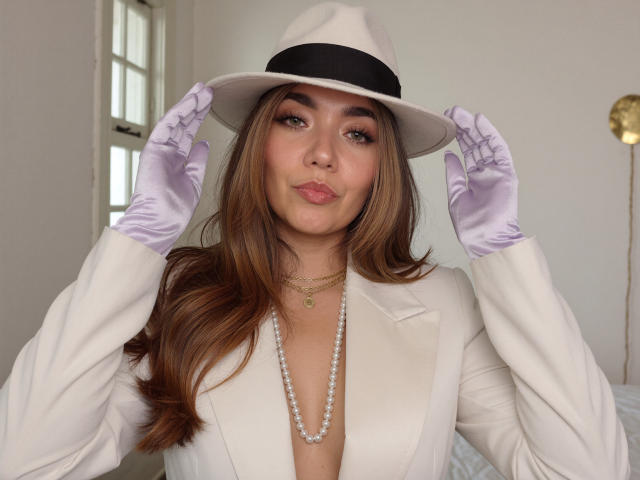 SofieWhite wearing a white hat, white jacket, purple gloves, pearl necklace, and gold necklace, with long brown hair, pouting, hands on hat. Boobs visible through jacket.