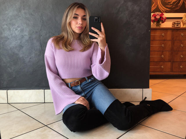 Sofie White mit blonden Haaren, einem rosa gestreiften Crop-Top, blauen Jeans, schwarzen Kniehochstiefeln, sitzend auf einem gefliesten Boden, die ein Spiegel-Selfie macht, ein schwarzes Handy haltend, süß, sexy, kokett..