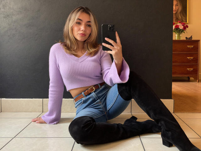 SofieWhite with blonde hair, purple ribbed crop top, blue jeans, black thigh-high boots, sitting on tiled floor, taking mirror selfie with phone, showing her tits.