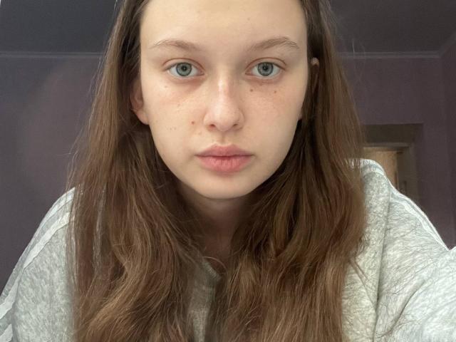 ClareVuturo heeft bleke huid, blauwe ogen en bruin golvend haar, draagt een grijze trui, heeft een neutrale uitdrukking en een kleine neusring aan de rechterkant..