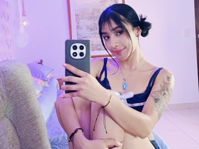 KatheLeclecr with dark hair in a bun, wearing a blue bra, holding a phone, showing off her tits, tattoo on left arm, sitting on a chair, smiling.