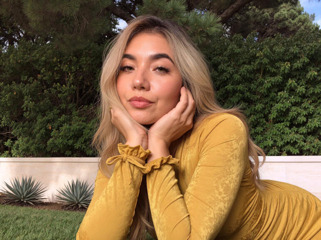 Sofie White tem cabelo loiro, lábios cheios e um rosto bonito, vestindo um vestido de veludo amarelo, com as mãos apoiadas nas bochechas, parecendo relaxada. Seus seios não estão visíveis..