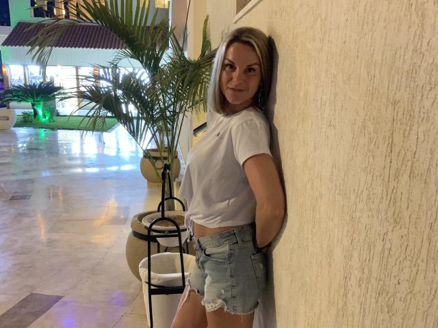 BelleLisaG with blonde hair, white t-shirt, and denim shorts, leaning against a wall, smiling, showing her ass in the image. She's standing next to a potted plant.