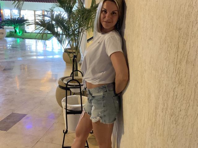 BelleLisaG leaning against a wall, wearing a white tee and frayed denim shorts, smiling with hands behind her back, blonde hair, standing on a tiled floor.