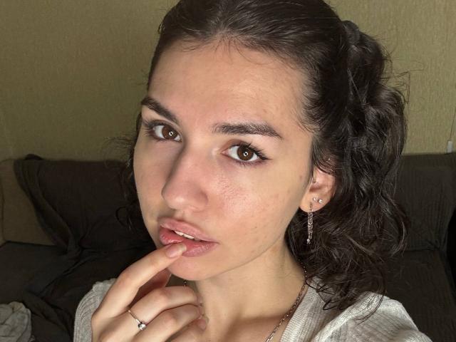 CristieTarvin with brown eyes, slightly parted pink lips, and finger touching them, has wavy dark hair, light skin, small earrings, and a silver ring on her left hand.