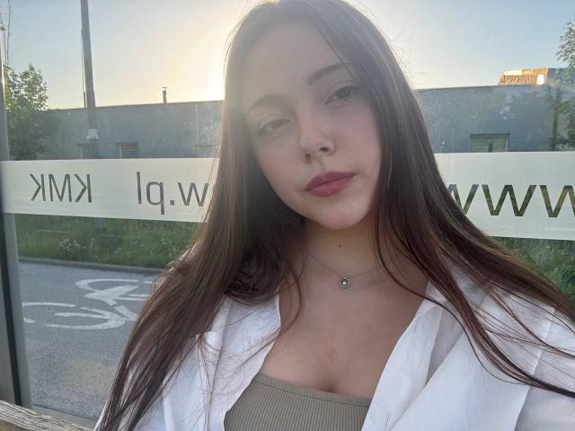Maida Cain, con i suoi lunghi capelli castani, indossa una camicia bianca sopra una maglia beige, ha un viso carino, un piccolo piercing al naso e una collana, ed è in piedi di fronte a una finestra di vetro..