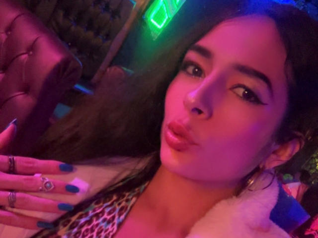 Ariel Nitzche, con los labios fruncidos, uñas azules, un piercing en la nariz y aros grandes, vestida con una blusa de estampado de leopardo, bajo luces de neón rosas y azules. Su mano tiene varios anillos..