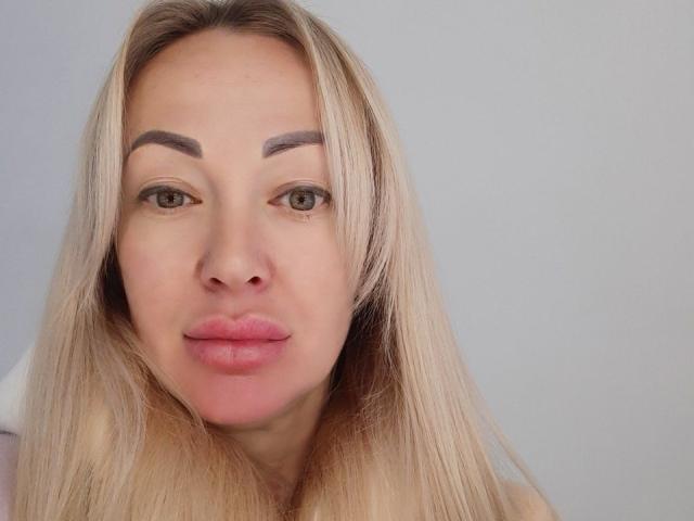 MysteryLaddy has blonde hair, thick eyebrows, green eyes, and pink lips, with a slight smile, looking directly at the camera. Her tits are not visible.