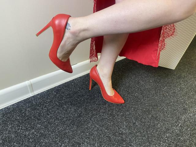 Aminas' pale legs in red high heels, one foot lifted, showing a small tattoo on her ankle. She's wearing a red lace dress, standing on dark carpet.
