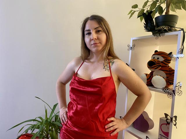 Aminas with medium tits, wearing a red satin dress, standing with hands on hips, brown hair, small tattoo on right shoulder, potted plants and tiger toy shelf in background.