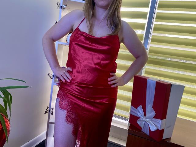 Aminas in a red satin slip with lace on the side, hands on hips, showing her ass. Gift box with white ribbon in the background.