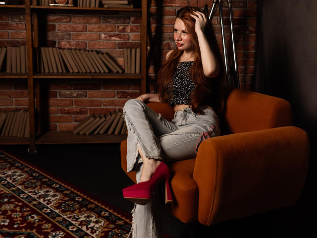AuroraSkye, avec de longs cheveux bruns, portant un haut noir à pois, un jean blanc effiloché et des talons rouges, assise sur une chaise orange, une main dans les cheveux, souriant..