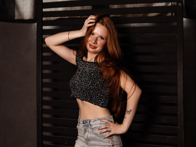 AuroraSkye, avec de longs cheveux roux, portant un crop top noir à pois et un jean bleu clair, la main dans les cheveux, souriante, montrant un tatouage sur son bras droit..