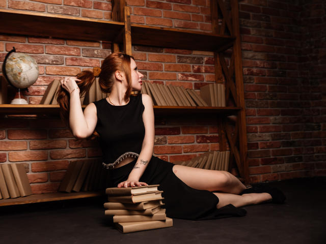AuroraSkye, con cabello rojo recogido en una coleta, está sentada en un suelo oscuro, vestida con un vestido negro sin mangas y zapatos negros, sosteniendo libros y un globo terráqueo, con una pared de ladrillos detrás..