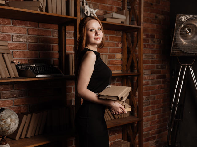AuroraSkye, avec des cheveux roux, une robe noire, tenant des livres, debout devant des étagères en bois, un mur de briques et un éclairage de studio, montrant légèrement ses seins et ses fesses en profil..