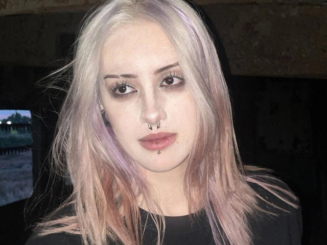Adina Tamburro, com cabelo rosa pálido e roxo, camisa preta, piercing no nariz, piercing no lábio e maquiagem discreta, olhando diretamente para a câmera em um ambiente com pouca luz..