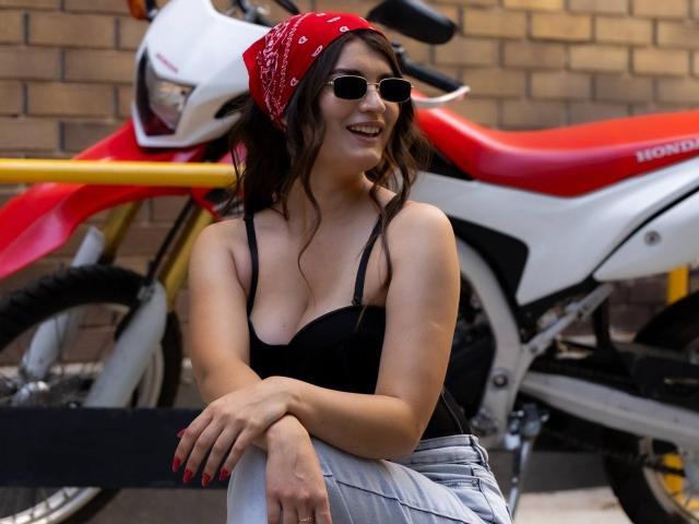 LauraLee69, com um lenço vermelho na cabeça, uma camiseta preta e calças cinzas, sentada em um suporte de bicicleta, sorrindo, usando óculos de sol, mostrando os seios, ao lado de uma motocicleta Honda vermelha e branca..