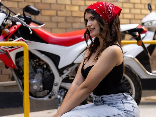 LauraLee69, com um lenço vermelho na cabeça, uma camiseta preta e calças jeans azuis claras, sentada em frente a uma motocicleta vermelha e branca, exibindo o bumbum e um sorriso sedutor..