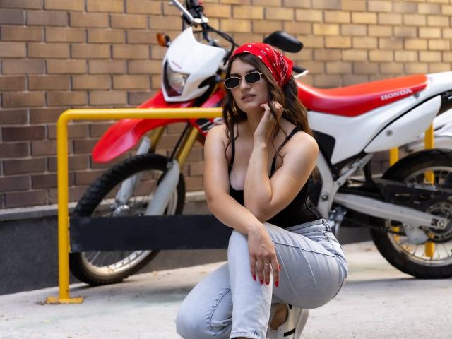 LauraLee69, avec de longs cheveux bruns, un bandana rouge, un débardeur noir laissant entrevoir de petites seins, un jean blanc et des lunettes de soleil, est accroupie près d'une moto Yamaha rouge et blanche..