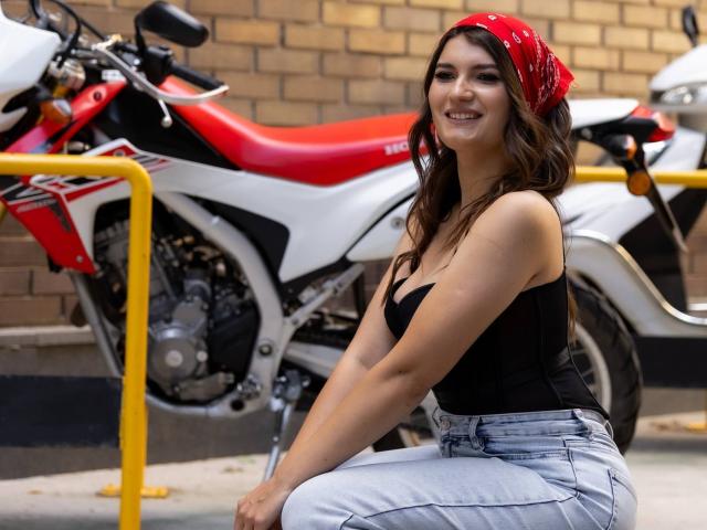 LauraLee69, com um lenço vermelho na cabeça, uma camiseta preta e calças cinzas, sentada em frente a uma motocicleta vermelha e branca, sorrindo, mostrando seus seios e nádegas, em um estacionamento..