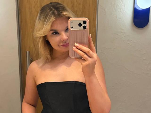 ChuHeal mit blonden Haaren, trägt ein schwarzes Top ohne Träger, hält ein rosa Handy, macht ein Selfie im Spiegel, lächelt leicht, zeigt ihre Brüste, im Badezimmer..