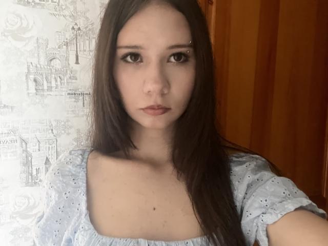 Lyndia Detlefs, com cabelos castanhos longos, olhos azul claros e uma pequena perfuração no nariz, vestindo uma blusa azul clara com bolinhas, de alças que deixam os ombros à mostra, revelando seu peito e seios pequenos..