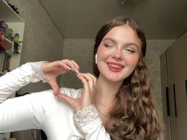 JulienneStradford with curly brown hair, white lace long-sleeve shirt, winking, pink lipstick, holding white earbuds, smiling, showing pink tongue, and making a heart shape with fingers.