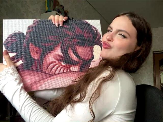Julienne Stradford, met lang bruin haar, een wit shirt met lange mouwen, houdt een pixelkunstwerk van een vrouwen gezicht vast, met samengeknepen lippen, en kijkt naar het kunstwerk, in een kamer..