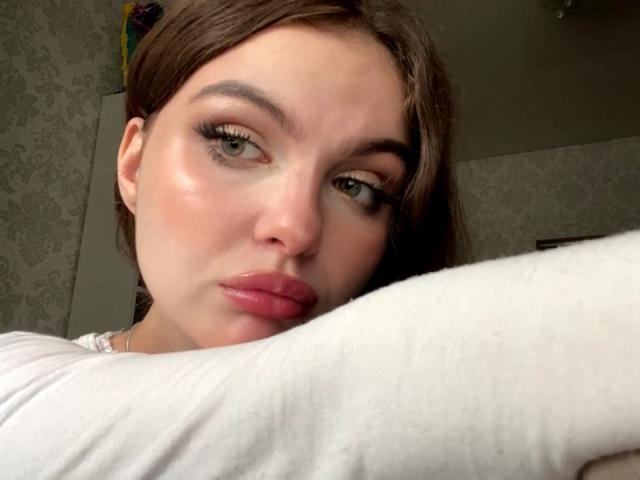 Julienne Stradford, avec ses lèvres roses et gonflées, ses yeux verts et ses cheveux bruns, a l'air légèrement agacée, elle porte une chemise blanche et son bras est visible au premier plan..