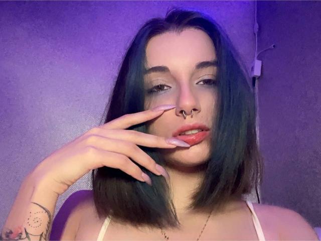 MackenzieCostantino with dark green hair, nose ring, and long nails, touching her red tongue, showing a small tattoo on her left wrist, wearing a white top.