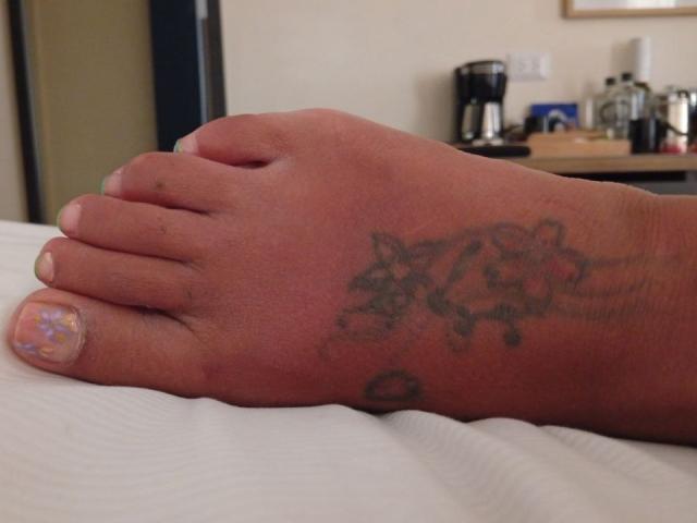 De voet van Laila Castillo heeft een zwarte tatoeage van bloemen en een klein hartje op haar enkel, en haar pinknagel is versierd met kleurrijk glitter. Haar tenen zijn lichtelijk vuil..