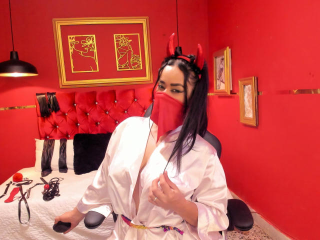 Anissa Ali, com cabelo preto, chifres vermelhos de diabo e máscara, vestindo uma túnica branca, expondo seus seios, sentada em um quarto vermelho com uma cama vermelha estofada e uma cadeira preta..