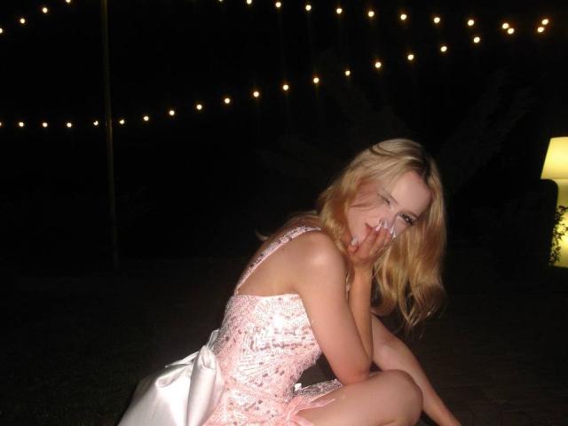 Dallia Banowski, rubia, con cabello ondulado, vestida con un vestido rosa con lentejuelas, sentada en el césped, con una mano en la mejilla, sonriendo, con una iluminación tenue gracias a luces de cadena, y una lámpara a la derecha..