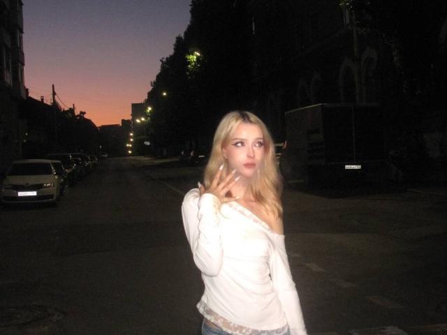 Blonde DalliaBanowski with puffy tits, off-shoulder white shirt, and slightly parted lips, flashing a peace sign at dusk in a dark, urban street.