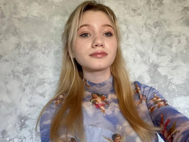 ElinorHamner with long blonde hair, wearing a sheer blue top with angel and cloud prints, has shiny pink lips and a slight smile, looking directly at the camera.