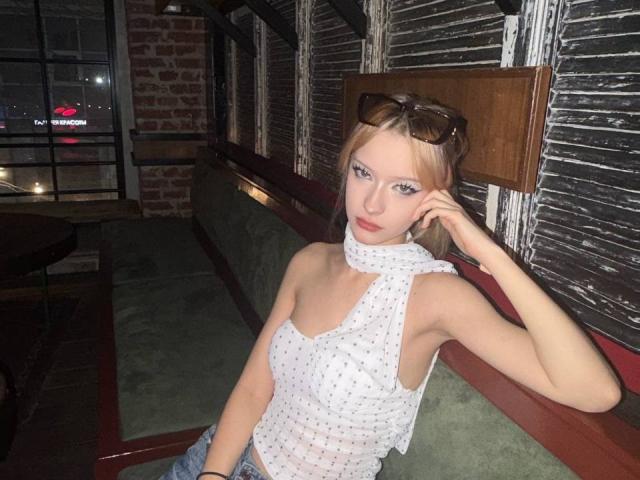 DalliaBanowski with small tits, wearing a white polka dot sleeveless top, sitting in a booth, hand in her blonde hair, sunglasses on her head, green cushioned seat.