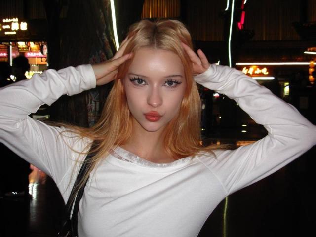 DalliaBanowski with blonde hair, pouty lips, and big tits, wearing a white long-sleeve shirt, hands in hair, in a dark, neon-lit urban setting, looking at camera.