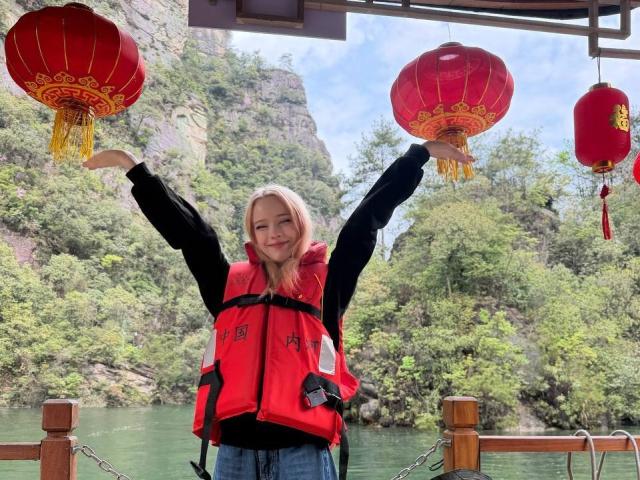 DalliaBanowski with arms raised, smiling, wearing a red life vest and black shirt, standing under red Chinese lanterns, blonde hair, green water, rocky hill, and forest in background.