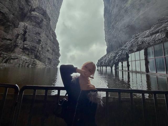 DalliaBanowski with blonde hair, wearing a black top, leaning against a metal railing in a rocky canyon with water, arms crossed, looking down, overcast sky, misty atmosphere.
