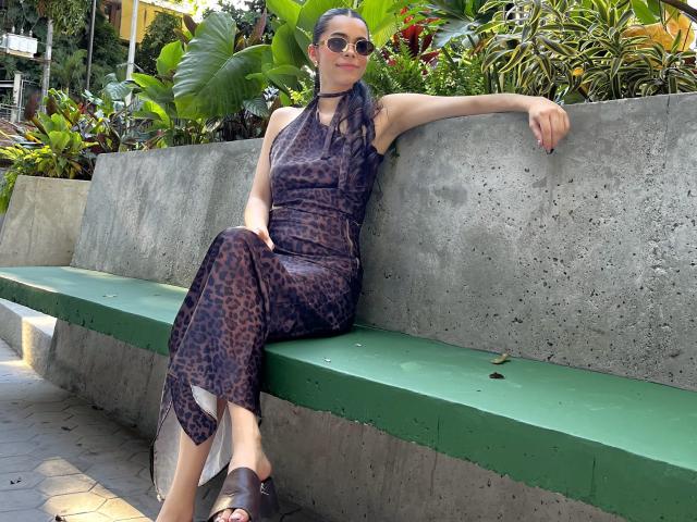 AlanaBlake, con capelli scuri, occhiali da sole e un vestito a stampa leopardo, seduta su una panca di cemento verde, con un braccio appoggiato sullo schienale, sandali marroni, piante verdi sullo sfondo, rilassata, sorridente, luce naturale..
