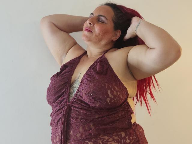 EmmaKings, con i capelli rossi, indossa un abito di pizzo viola scuro, ha le braccia dietro la testa, mostrando il suo seno prosperoso e un tatuaggio sul fianco. Sta sorridendo..