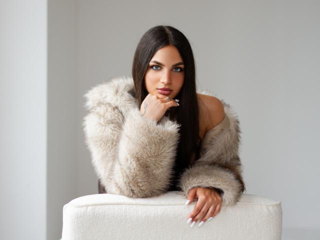 CarlaXQueen mit langen, braunen Haaren, blauen Augen und vollen Lippen, trägt einen flauschigen, beigefarbenen Fellmantel, lehnt sich an ein weißes Kissen und blickt mit ernstem Gesichtsausdruck direkt in die Kamera..